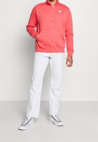 Korallfärgad Nike-tröja med halvdragkedja, känguruficka och ribbade muddar, kombinerad med vita jeans med raka ben och gråa sneakers.