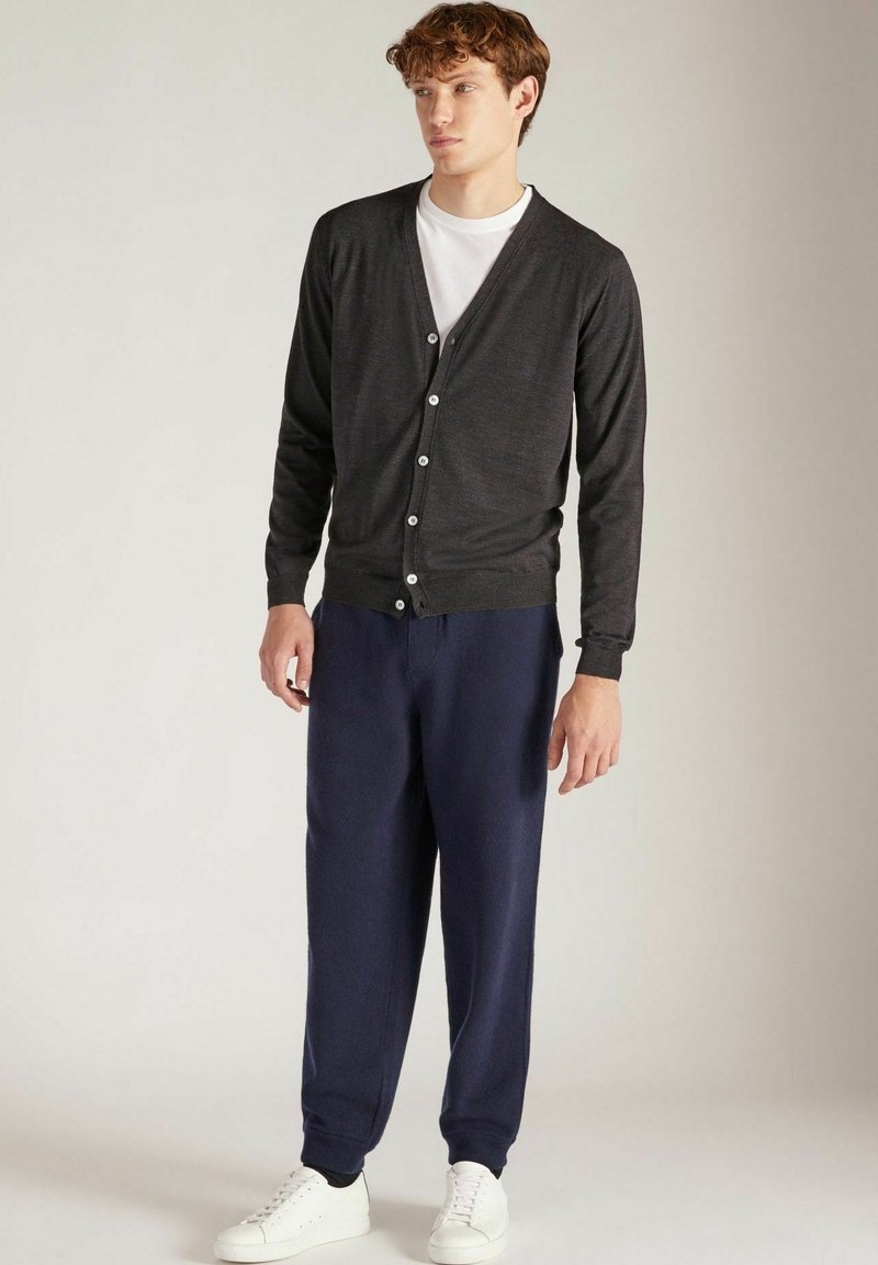 Donkergrijze cardigan met een V-hals, knopen aan de voorkant, gedragen over een wit T-shirt, gecombineerd met navy joggers en witte sneakers.