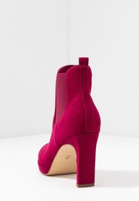 Botín de ante fucsia con tacón, paneles laterales elásticos, punta redonda y superficie texturizada. Cuenta con un tacón resistente y una tira para facilitar el calzado.