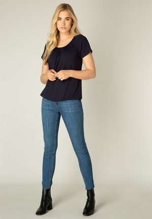 Marineblauwe top met korte mouwen en plooidetail; gecombineerd met slim-fit blauwe jeans en zwarte enkellaarsjes. Stevige stof, ontspannen silhouet.