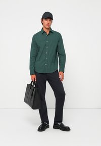 Tânăr îmbrăcat cu o cămașă verde cu model, pantaloni negri, șapcă neagră și pantofi negri, ținând o geantă de mână din piele neagră, pe un fundal alb.