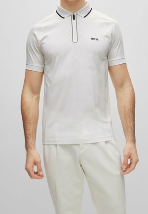 Homme portant un polo blanc à manches courtes avec une fermeture éclair bordée de noir et le logo "BOSS" sur la poitrine gauche, porté avec un pantalon blanc.