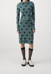 Grünes gestricktes Zweiteiler-Outfit mit marineblauen Herzmustern. Langärmliges Oberteil mit rundem Halsausschnitt und eng anliegender Rock. Kombiniert mit schwarzen knielangen Strümpfen und Slippern.