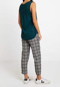 Kvinna i ärmlös turkos blus, svart och vit rutiga byxor, och vita sneakers som står mot en enfärgad vit bakgrund.