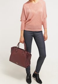 Borsa color bordeaux con parte frontale liscia e lato texturizzato, abbinata a un maglione lavorato a maglia rosa chiaro e pantaloni grigi aderenti.