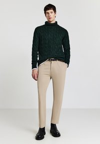 Grüner kabelgestrickter Rollkragenpullover mit strukturiertem Muster, kombiniert mit beigen Slim-Fit-Hosen und schwarzen Knöchelstiefeln für einen geschichteten Look.