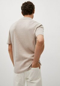 Polo beige à manches courtes avec un tricot texturé, présentant un col et une coupe droite. Porté avec un pantalon blanc.