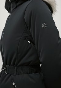 Sorte isolerede jakke med hætte i pels, elastisk taljebælte, sidelomme med lynlås og et broderet snefnuglogo på ærmet.