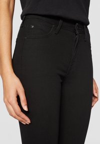 Jean en denim noir avec une coupe slim, des coutures contrastantes, des poches avant et une fermeture éclair, avec une main placée sur la cuisse.