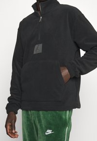 Pullover in pile nero con zip a quarto, tasca kenguru e patch con logo sul petto. Presenta polsini a costine e una vestibilità comoda.