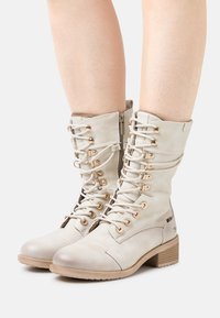 Lace-up ankle boots in soft light beige leather with a rounded toe, gold hardware, side zip closure, and a stacked heel.