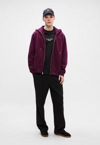 Bordo kapucis ar rāvējslēdzēju un priekšējām kabatām, valkāts virs melnas t-krekliņa ar baltu tekstu. Saskaņots ar brīviem melniem biksēm un melnām kurpēm.