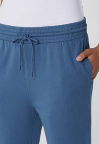 Blaue Jogginghosen mit elastischem Bund und Kordelzug, getragen mit einem in die Hose gesteckten weißen Hemd, die Hand ruht in der rechten Tasche.