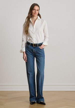 Kvinde stående med langt brunt hår, iført en hvid skjorte med knapper, blå jeans med sort bælte og sorte spidse sko.