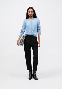 Femeie în picioare, purtând un cardigan bleu deschis, pantaloni negri cu talie înaltă, cizme negre până la gleznă, ținând o geantă de umăr din piele de șarpe gri.