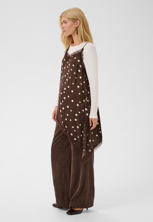 Femme en haut blanc à manches longues, robe nuisette marron à pois, pantalon marron large et chaussures noires, debout de profil.