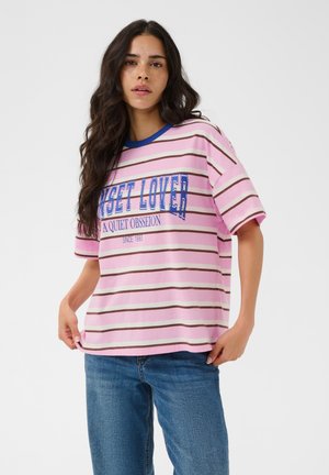 Femme aux longs cheveux foncés portant une chemise rayée rose et marron avec le texte "Sunset Lover" et un jean bleu sur un fond uni.