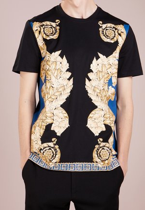 Zwarte korte mouwen shirt met gouden barokke bladsieraden en blauwe geometrische rand, gedragen door een persoon met handen in de zakken.