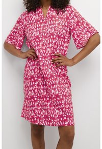 Kvinna med lockigt hår som bär en rosa och vit mönstrad knä-lång klänning med korta ärmar, stående med händerna på höfterna.