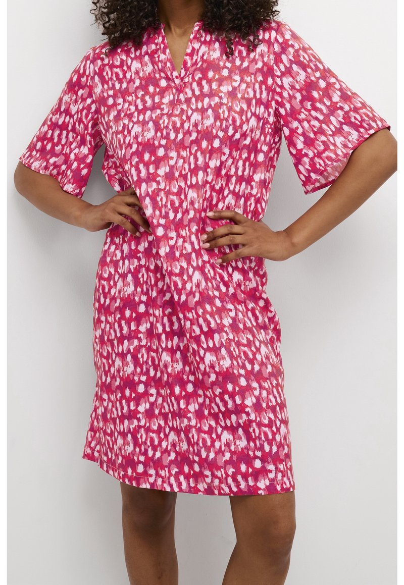 Kvinna med lockigt hår som bär en rosa och vit mönstrad knä-lång klänning med korta ärmar, stående med händerna på höfterna.