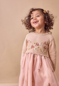 Helles rosa gestricktes Sweater mit floralem Stickerei am Saum, dazu ein fließendes durchsichtiges rosa Kleid, das mit bestickten Blumen verziert ist.