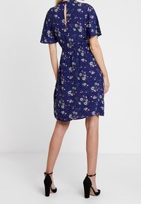 Marinblå blommig klänning med korta ärmar, åtsittande midja och en nyckelhålsdetalj på ryggen. Svarta klackar kompletterar outfiten.