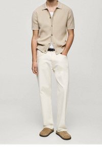 Cardigan beige a coste con maniche corte, t-shirt bianca sotto, abbinato a pantaloni dritti color avorio e scarpe slip-on marrone chiaro.