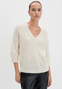 Weibliches Model trägt einen weißen strukturierten V-Ausschnitt-Pullover mit drei Viertelärmeln und schwarze Lederhosen vor einem schlichten Hintergrund.