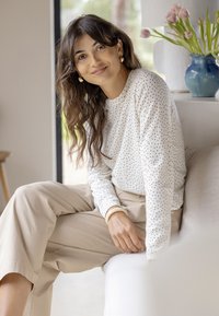 Femme aux cheveux bruns ondulés portant un pull blanc à pois et un pantalon beige, assise sur un canapé, souriante, avec un vase bleu de fleurs à proximité.