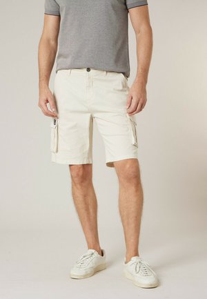 Man die beige cargoshorts met zijzakken, een grijs T-shirt en witte sneakers draagt, staand op een beige achtergrond.