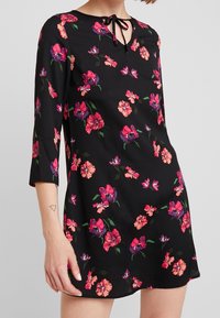 Schwarzes Kleid mit Dreiviertelärmeln, das mit floralen Mustern in Pink, Rot und Lila versehen ist. Schlüsselbeen- Ausschnitt mit einem Bindedetail. Glatte Textur.