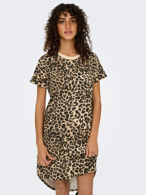 Kvinde med krøller iført en kortærmet kjole med leopardprint, stående mod en lys baggrund.