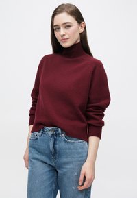 Bordowy sweter z dzianiny z golfem i luźnymi rękawami, zestawiony z jasnoniebieskimi, znoszonymi dżinsami. Miękka faktura, prążkowany kołnierz i wykończenia przy mankietach.