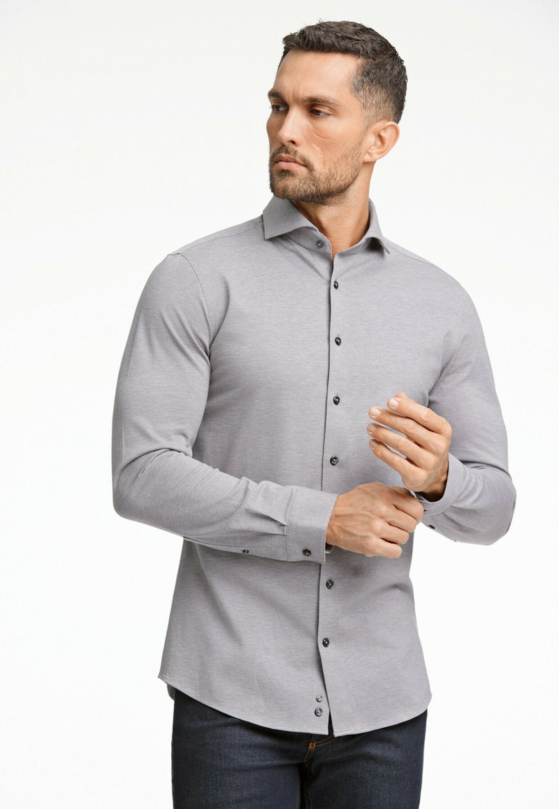 Grijze langdraagshirt met een knoopsluiting, zwarte knopen en een getailleerde pasvorm, gecombineerd met donkere spijkerbroek. Glad stoftextuur.