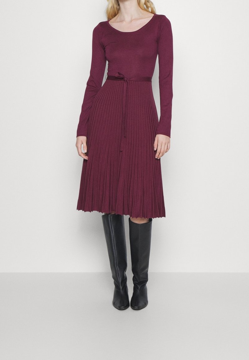 Robe prune ajustée à manches longues avec une jupe plissée et une ceinture nouée intégrée. Associée à des bottes noires élégantes montant jusqu'au genou.