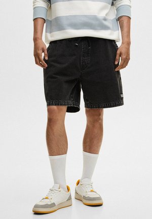 Czarne szorty jeansowe o luźnym kroju, z postrzępionym dołem i bocznymi kieszeniami, zestawione z białymi skarpetkami i sneakersami z żółtymi akcentami.