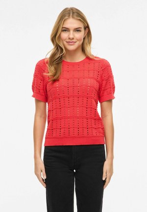 Jauna moteris su blondiniais plaukais, dėvinti raudoną trumpomis rankovėmis megztą viršutinį drabužį su akių raštais ir juodas kelnes, stovinti priešais baltą foną.