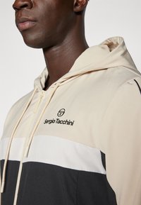 Sergio Tacchini hættetrøje med lynlås i beige, med sorte og hvide farveblokke, tekstureret stof og logo på brystet nær lynlåsen.