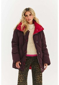 Pikowana bordowa kurtka z jasnoczerwonym kołnierzem, z przeszywanym wzorem i dużymi guzikami, noszona na kremowym swetrze i spodniach w panterkę.