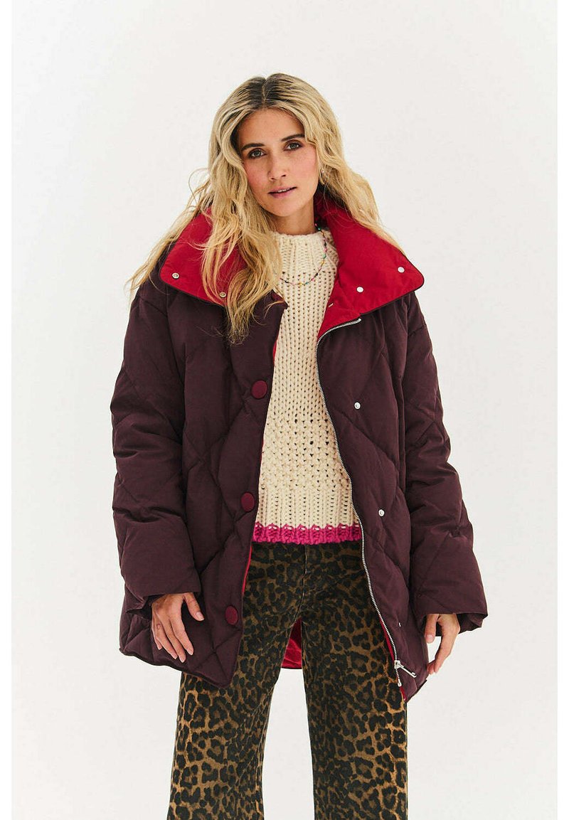 Pikowana bordowa kurtka z jasnoczerwonym kołnierzem, z przeszywanym wzorem i dużymi guzikami, noszona na kremowym swetrze i spodniach w panterkę.
