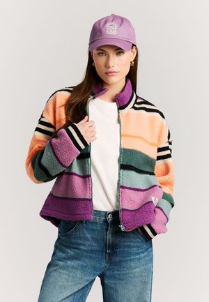 Vrouw met een meerkleurige gestreepte fleecejas, wit overhemd, blauwe spijkerbroek en paarse pet met de tekst "Good Vibes Protest" en zonnegrafiek.