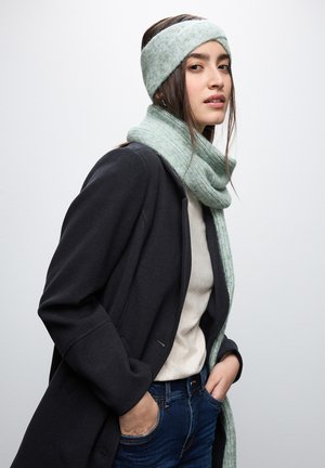 Mintgrøn strikket tørklæde og pandebånd båret med en sort frakke og beige top, der har en blød stofstruktur og en snug pasform.