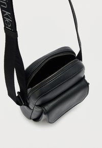 Μαύρο δερμάτινο τσάντα χιαστί με υφή, κλεισίματα με φερμουάρ και ρυθμιζόμενο λουρί. Συμπαγής σχεδίαση με μία κύρια θήκη και μια μικρή μπροστινή τσέπη.