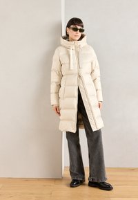 Beige gewatteerde jas met capuchon, ritssluiting en koord. Lange mouwen, gebandeerde structuur. Gecombineerd met donkergrijze wijde jeans en zwarte schoenen.