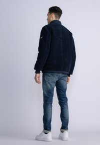 Navy blue fleece jacket with a textured finish, ribbed cuffs, and a stand-up collar, paired with light blue jeans and white sneakers.