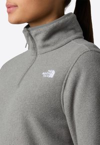 Pullover grigio in pile con zip a quarti, caratterizzato da un colletto alto e un logo ricamato sul petto. Il tessuto ha una finitura morbida e testurizzata.