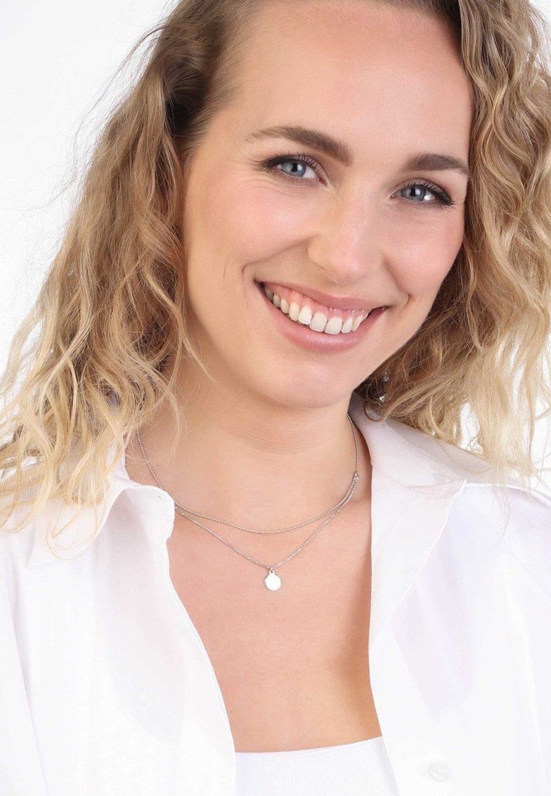 Zilveren gelaagde halsketting met een kleine ronde hanger. Model met golvend, blond haar draagt een witte blouse tegen een witte achtergrond.
