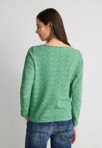 Grüner Strickpullover mit langen Ärmeln und weitem Ausschnitt, der ein strukturiertes Muster aufweist. Getragen mit blauen Jeans, die eine lockere Passform zeigen.
