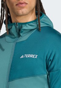 Teal Terrex jacket with a zippered front, hood, and contrasting fabric panels. Features embroidered logo and smooth, textured fabric.