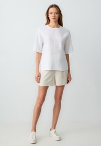 T-shirt bianca in lino con maniche corte e larghe, scollatura rotonda, indossata con pantaloni corti beige e sneakers bianche, su uno sfondo neutro.